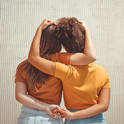 Strong female friendship. Rear view two teen girls best friends holding hands behind back and hugging while standing in front of beige wall outdoors